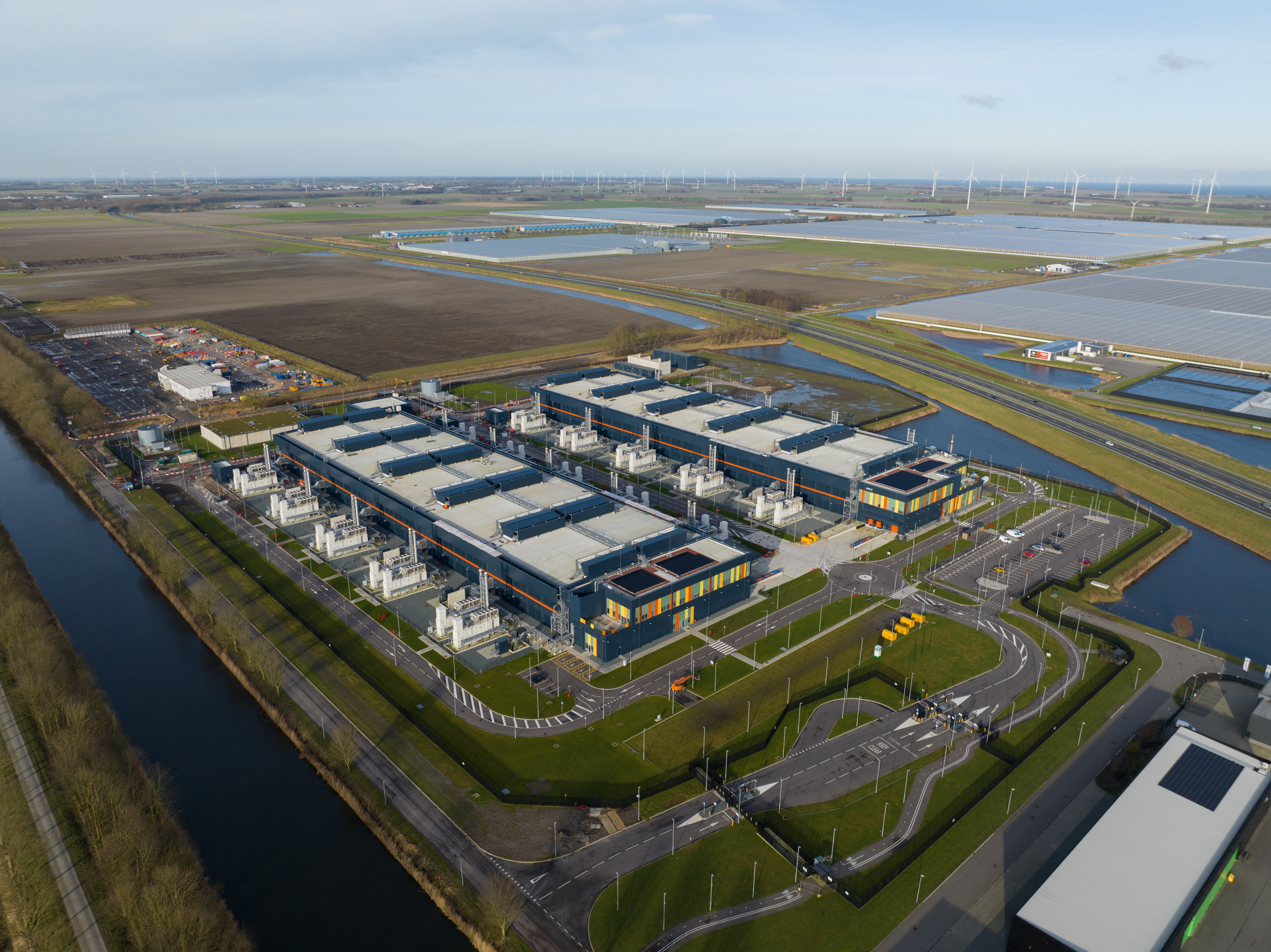 Aerial view of a large data center with multiple buildings, energy infrastructure, water channels, and surrounding solar and wind energy systems.