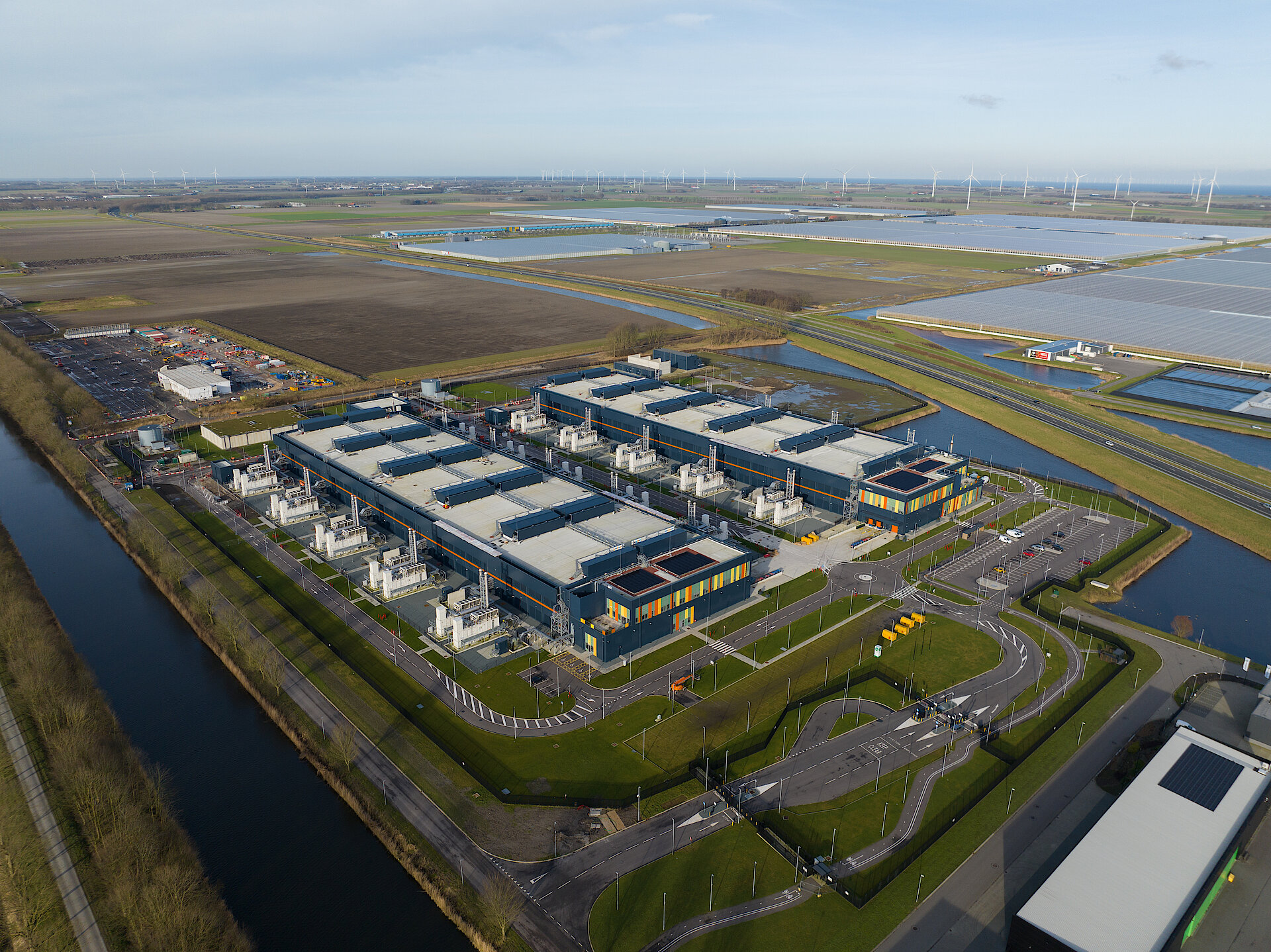 Aerial view of a large data center with multiple buildings, energy infrastructure, water channels, and surrounding solar and wind energy systems.