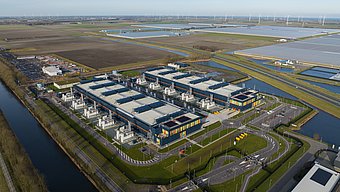 Aerial view of a large data center with multiple buildings, energy infrastructure, water channels, and surrounding solar and wind energy systems.