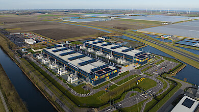 Aerial view of a large data center with multiple buildings, energy infrastructure, water channels, and surrounding solar and wind energy systems.